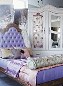 Ein antikes Polsterbett mit Seidenkissen vor einem romantischen, weissen Kleiderschrank mit Spiegeltüren