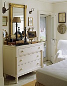 White chest of drawers in bedroom below gilt-framed mirror