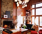 Wohnzimmer im Landhausstil mit offenem Kamin in der Natursteinwand; eine hohe Fensterfront sorgt für helles Licht im ansonsten holzverkleideten Raum
