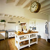 Große, weiße Küche mit Balkendecke, einer Bahnhofsuhr und einem zentralen Küchenblock in einfachem, ländlichen Stil