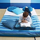 Frau liegt lesend auf Matratze mit blauen Kissen und blauer Decke