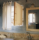 Ausschnitt eines Badezimmers mit Granitwaschtisch und barockem Wandspiegel neben einem kleinen Fenster mit Holzklappe
