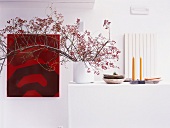 Ein rotes Gemälde und Blumenzweige in einer Vase neben Schalen und Kerzen in einem schneeweissen Raum