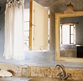 Ausschnitt eines Badezimmers mit Granitwaschtisch und barockem Wandspiegel neben einem kleinen Fenster mit Holzklappe
