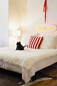 Eine schwarze Katze auf einem poppigen Bett mit weisser Felldecke und -kissen