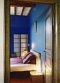 Blick durch eine rustikale Holztür ins blaue Schlafzimmer mit Holzbalkendecke