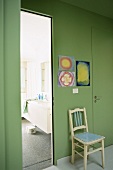 Ein antiker Stuhl unter Kinderzeichnungen an einer grünen Wand, daneben das Badezimmer