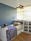 Weisses Schubladenbett vor der blauen Wand eines Kinderzimmers mit Holzdecke und Fensterfront