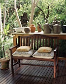 Holzbank mit Sitzkissen und ein Tisch mit alten Eisengieskannen auf einer Holzveranda im Grünen