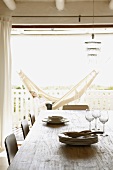 Rustikaler Holztisch mit Weingläsern und Tellern vor Frau in Hängematte auf der Terrasse