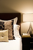 Bett mit verschiedenen Zierkissen und Nachttisch mit Lampe & Büchern im Schlafzimmer (Ausschnitt)