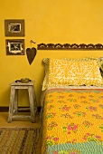 Bett mit gemusterter Tagesdecke und rustikalem Hocker vor gelber Wand