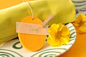 Grüne Stoffserviette mit Namensschild und gelbe Blumenblüte auf Teller dekoriert