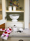 A white crockery cupboard with an open door and an old enamel colander inside