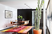 A dining table with cantilever chairs on a striped rug on black floor tiles with a cactus in an orange pot