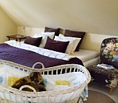 A bed with a purple throw under a slopping roof and a cat in a Moses basket