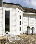 A front door and a floor-to-ceiling window in a white wooden house with garden chairs and plant pots outside