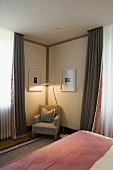 Light gray armchair with a floor lamp in the corner of a bedroom and floor length curtains at the window