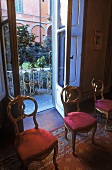 Antique wooden chairs upholstered in red in front of an open terrace door with a view of a garden