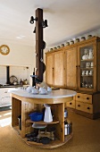 A kitchen in a country house featuring a wooden counter island with a marble work surface built around a wooden pillar hung with spotlights in front of cupboards
