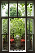 A view through an open window onto a wooden terrace with red garden chairs and a table