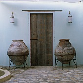 A Mediterranean courtyard - heavy amphorae in metal stands in front of a rustic wooden door on a natural stone floor