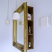 A bathroom corner - a mirror in a gold frame in front of a shelf with side opening and a pendant lamp with white glass shades