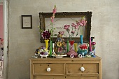 A colourful collection of vases on a wooden chest of drawers and a picture frame on a grey wall
