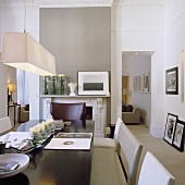 A dining area with a pendant lamp and a white shade in front of a grey wall with a fireplace and a view through a doorway