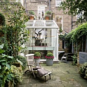 Two story greenhouse in front of a home with brick facade and old wooden deckchair in a backyard