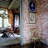 A country-style chair against a patterned wall paper in a hallway with a view through an open door into a living room