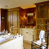 A bathroom in an English country house with a free-standing bathtub in front of wooden, floor-to-ceiling cupboards and a washstand against a red illuminated wall