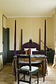 A bedroom - a old fashioned chair and table in front of a four poster bed with pillars
