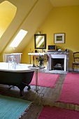 A yellow-painted attic bathroom with a fireplace and pink bathmats in front of an antique, free-standing bathtub