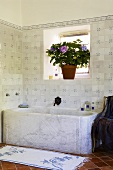 A stone bathtub against a tiled wall with a window and a pot of flowers on the window sill
