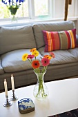 Flowers in a vase and candlesticks on a coffee table in from of a gray sofa with striped pillows