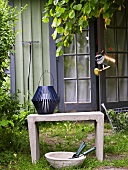 A blue lantern on a stone bench and bowl of garden tools in front of a garden shed