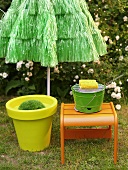 Yellow plastic plant pot and orange stool in front of green sun umbrella made from bast