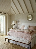 A detail of a traditional bedroom decorated in neutral colours, pattern wallpaper, beamed ceiling, double bed with upholstered headboard, bench seat, lamp, mirror, natural flooring,