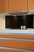 Küchenschrankausschnitt mit orangefarbenen Fronten und Edelstahltopf vor schwarzer Rückwand