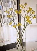 A detail of an arrangement of yellow flowers in a glass vase, black and white print in frame placed on mantelpiece,