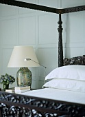 A detail of a traditional bedroom with painted panelling, carved wooden four poster bed, bedside table, lamp