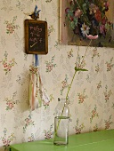A flower in a glass bottle in front of a wall hung with floral paper