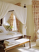 Schlafzimmer mit Himmelbett mit weissen Vorhängen, Toile-de-Jouy Vorhängen am Fenster und mit einer weissgepolsterten Sitzbank am Ende des Betts