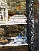 Plates, a bread basket, tablecloths and a bird cage on a shelf against a brick wall