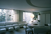 Weisses Sofa vor dem Fenster und runder Steintisch mit weissen Leder-Sitzbänken in einem offenen Wohn- und Esszimmer