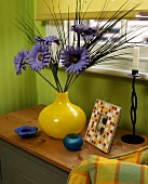 Gelbe Vase mit violetten Plastikblumen auf Holzablage vor grüner Wand