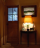 Gemälde über kleinem Konsolentisch mit brennender Lampe neben halbverglaster Zimmertür mit Blick in Küche