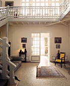Freitreppe und umlaufende Galerie in lichtdurchfluteter Eingangshalle mit hellblauem Teppich vor gelb gepolstertem, antikem Sessel auf Mosaikboden