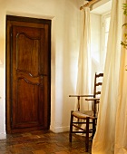 Alte Mahagoni-Tür neben antikem Holzstuhl mit Sitzgeflecht vor Fenster mit hellen Vorhängen